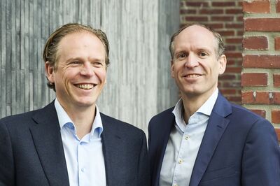 Two smiling Actipro executives in dark blazers against a grey wood and brick wall