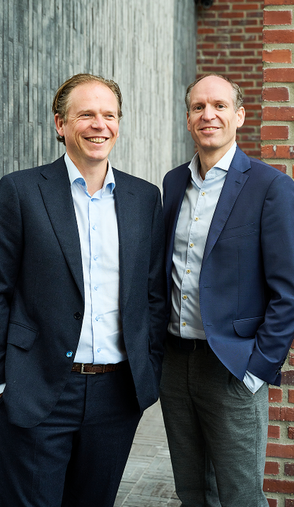 Two smiling businessmen in dark suits standing against a grey wood and brick wall