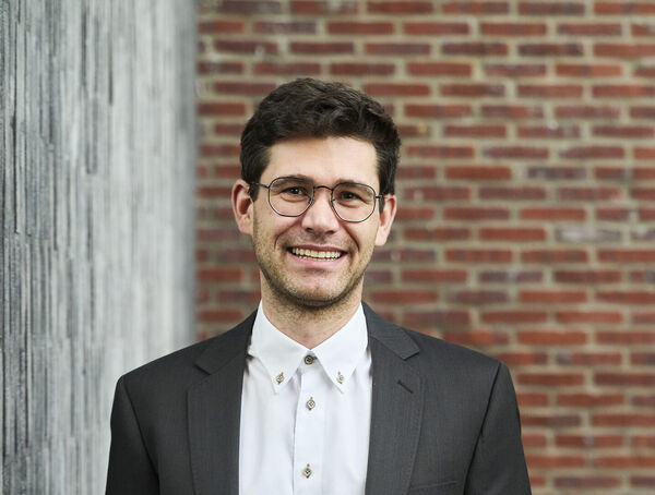 Smiling team member with glasses in a dark suit jacket and white shirt in front of a brick wall