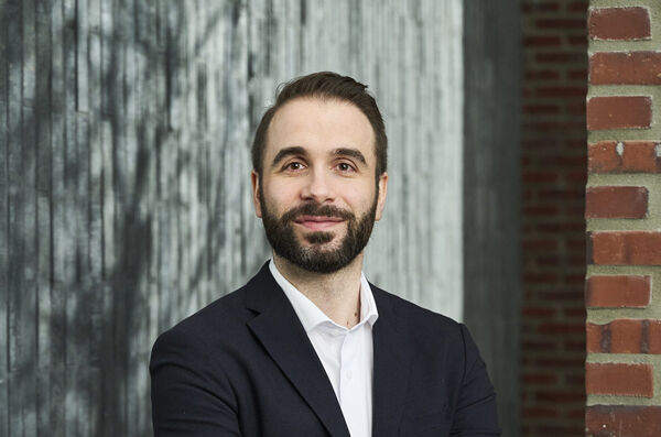 Bearded team member in a navy blazer and white shirt in front of a concrete and brick wall