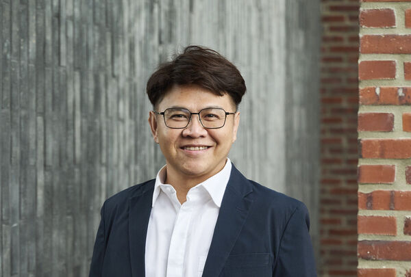 Smiling team member with glasses in a navy jacket and white shirt in front of a concrete wall