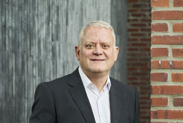 Grey-haired team member in a dark suit jacket and white shirt in front of a brick wall